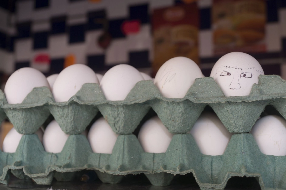 Cartón verde de huevos blancos donde destaca uno en la esquina superior derecha que tiene un rostro dibujado a lápiz.